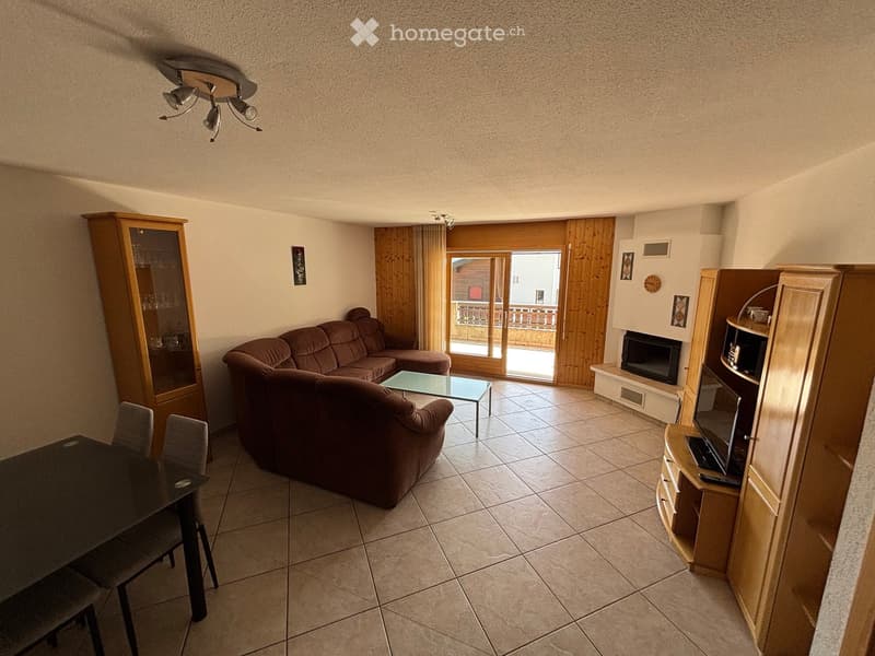 Living room with a brown sectional sofa, glass coffee table, dining table with chairs, wooden cabinet, fireplace, and TV stand. Large sliding glass door leads to a balcony.