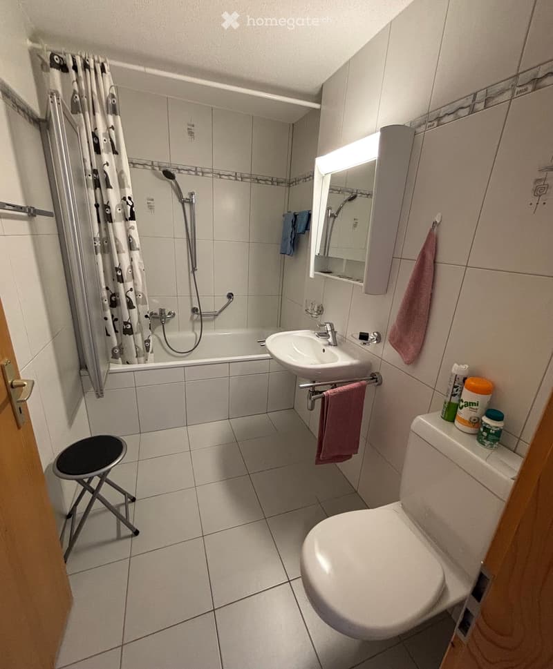 A compact bathroom with a bathtub, sink, toilet, and a small black stool. The space features tiled walls and floor, a shower curtain, and a mirror above the sink.