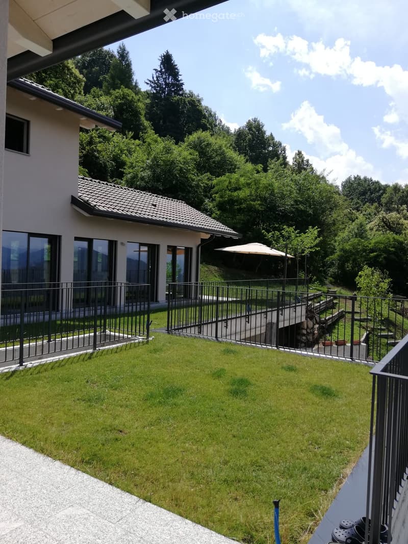 modern house with glass walls, balcony, green grass lawn, metal railings, raised ground floor, view of mountains and trees
