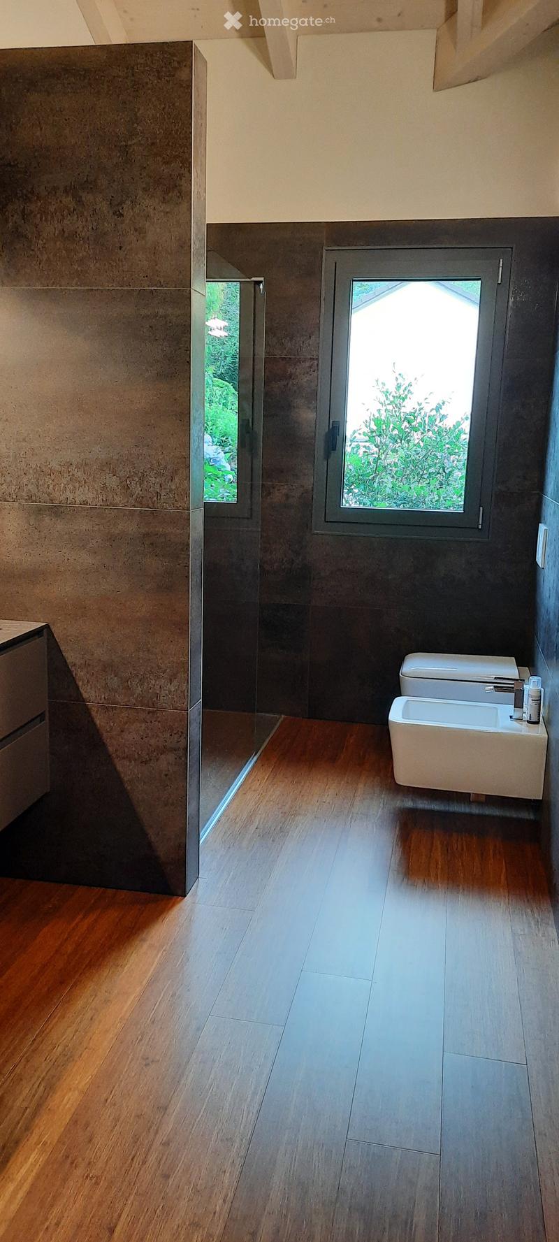 A modern bathroom with wooden floor, stone walls, glass partition, white sinks, and a window