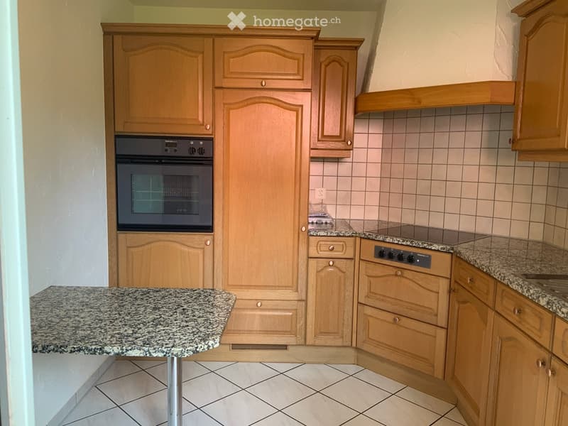 A modern kitchen with light wooden cabinets, granite countertops, and built-in appliances including an oven and stove.