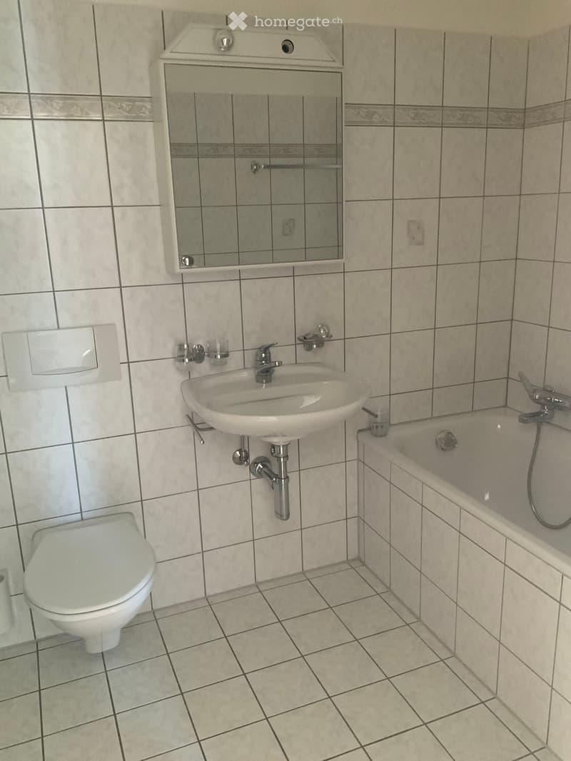 Bathroom with a white sink, toilet, bathtub, and a mirror above the sink. The walls and floor are tiled with light-colored tiles.
