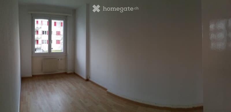 Empty room with wooden flooring, white walls, and a window showing a view of a building with red accents.