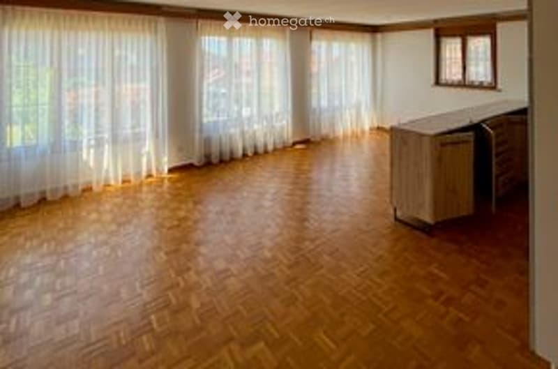 Empty room with wooden flooring, large windows with white curtains, and a small cabinet in the corner.