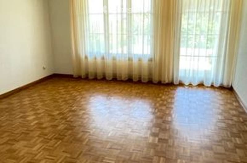 Empty room with wooden parquet flooring, large windows with white curtains, and white walls.