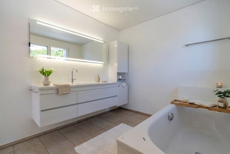 Modern bathroom with a large bathtub, white vanity with sink and mirror, and decorative items including a vase with white flowers, candles, and a plant on a wooden tray.