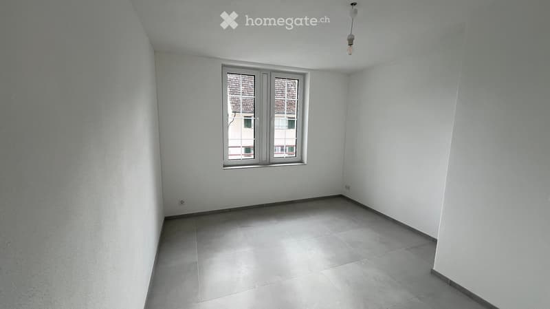 Empty room with white walls, tiled floor, and a single window with a view of a neighboring building.