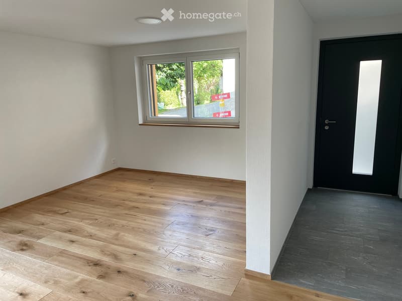 Empty room with light wooden flooring, white walls, a large window with a view of greenery, and a black door.