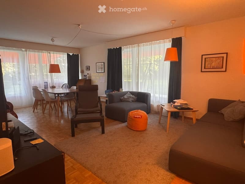 A modern living room with a sofa, a chair, a small table, a round stool, a dining table with chairs, and a TV stand with a screen. The room has large windows with curtains and is well-lit with hanging lamps.