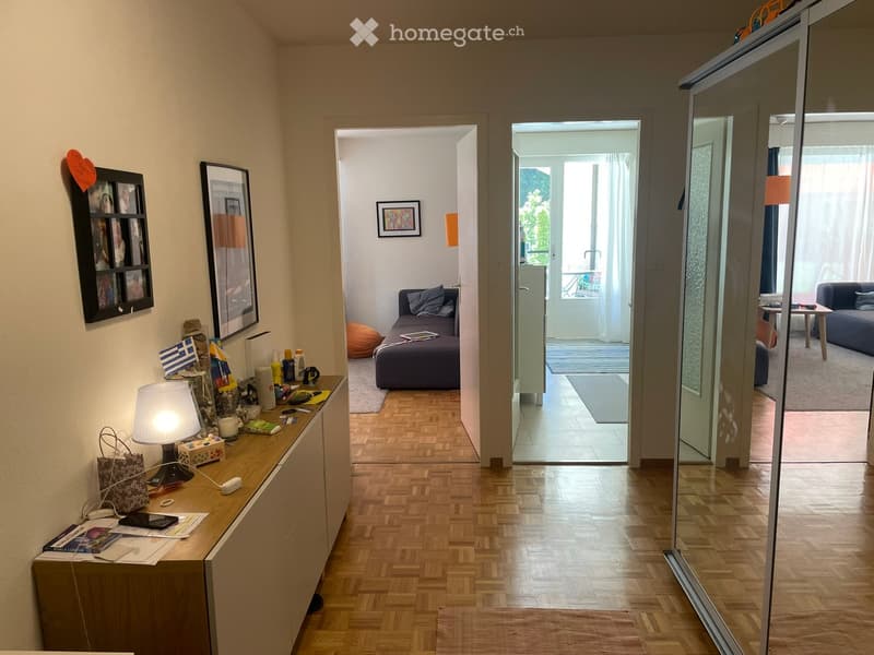A modern, well-lit hallway with wooden flooring, leading to a living area with a sofa, ottoman, and a small table. The hallway features a wooden side table with a lamp, picture frames, and various small items.