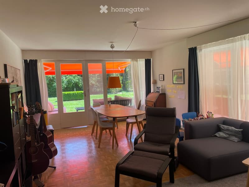A modern living room with a dark grey sofa, a wooden dining table with chairs, a guitar, and a comfortable chair with an ottoman. Large windows with curtains lead to a garden view.