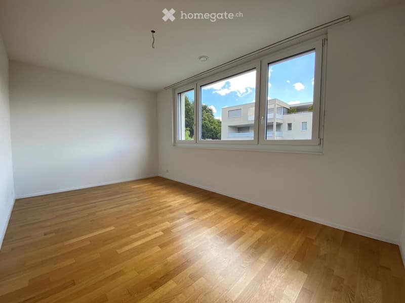 Empty room with wooden flooring and large windows offering a view of a building and trees outside.