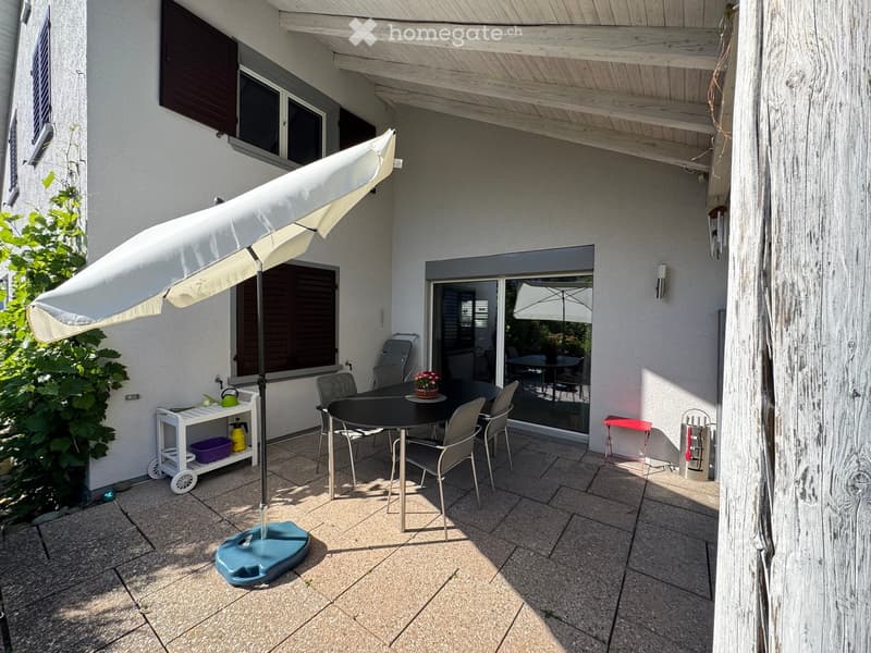 A covered outdoor patio with a dining table, chairs, umbrella, and a small cart with items.