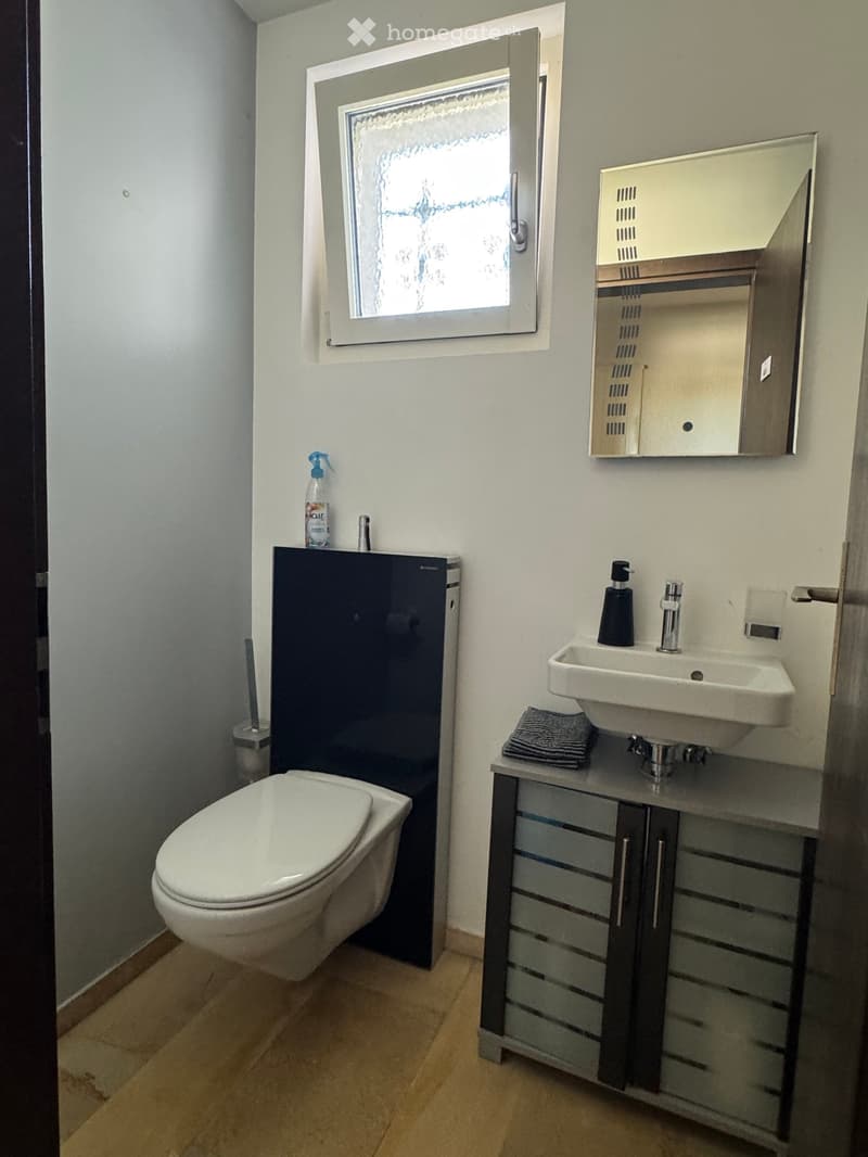 Modern bathroom with a white toilet, sink, and mirrored cabinet. Features a window with frosted glass and a small towel on the sink counter.