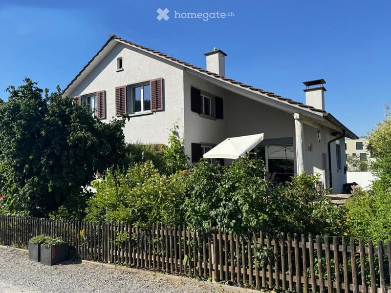 Two-story house with white exterior, brown shutters, and a covered patio area with an umbrella. The house is surrounded by a wooden fence and lush greenery.