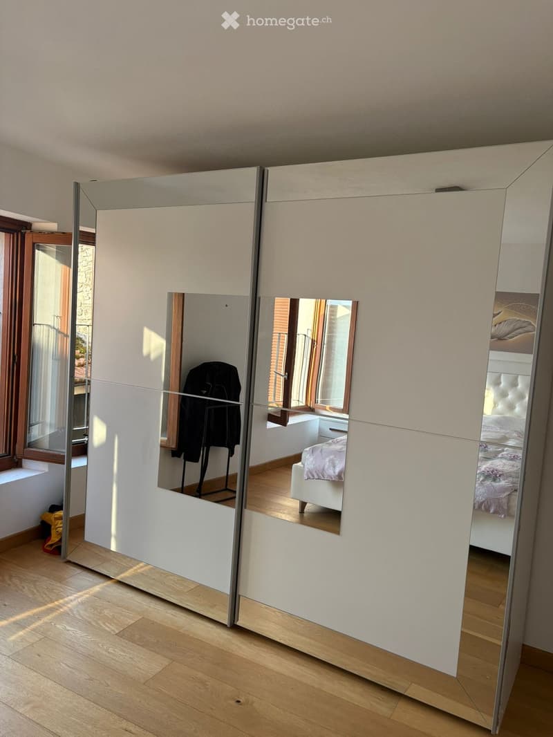 Modern bedroom with large mirrored wardrobe, wooden floor, and visible bed and nightstand through the open wardrobe doors.