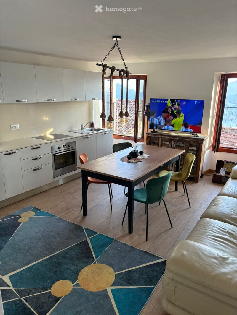 Open-plan living and dining area with a sofa, dining table, chairs, television, kitchen appliances, and a view of the mountains through large windows.