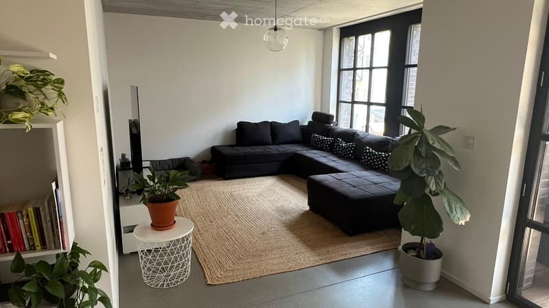 Modern living room with a dark sectional sofa, a small round white table with a potted plant, a bookshelf with books and a TV, and large windows letting in natural light.