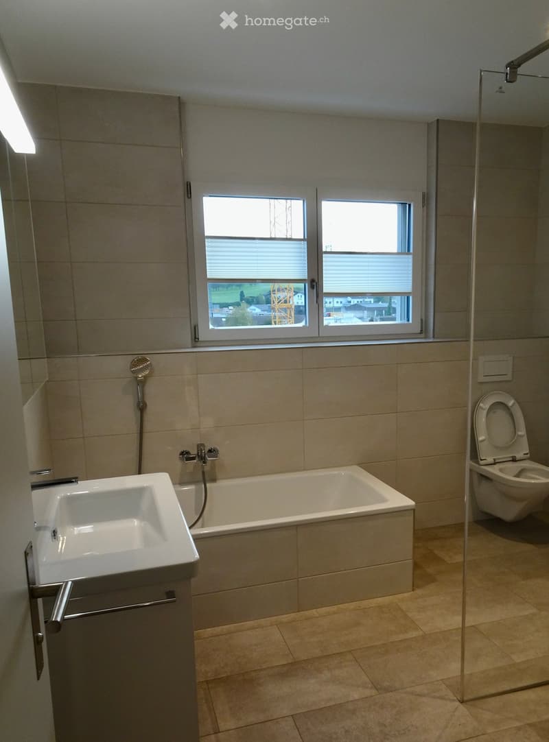 Modern bathroom with a bathtub, toilet, sink, and showerhead. Features large windows with blinds and tiled walls and floor.