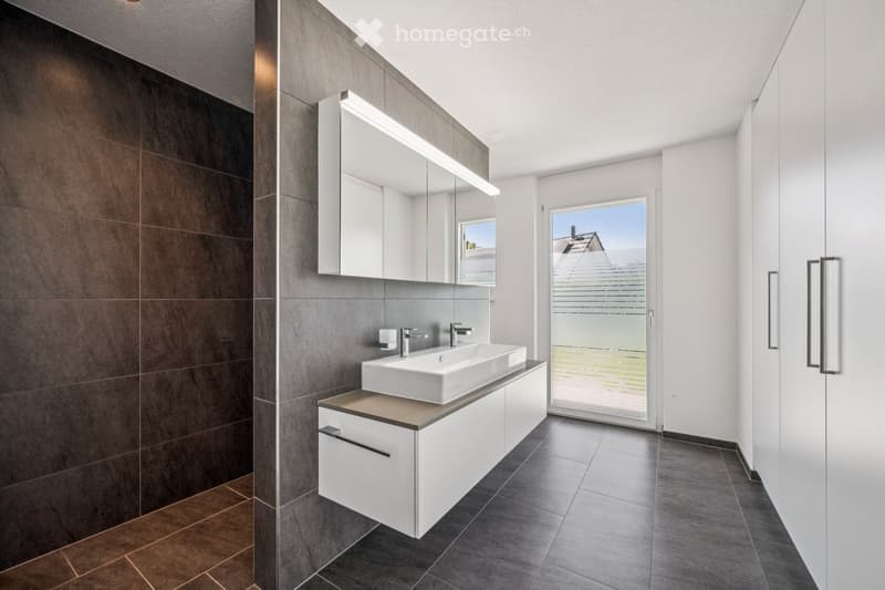 Modern bathroom with a white sink, mirrored cabinet, and dark tiled walls. Features a large window offering a view of the exterior.