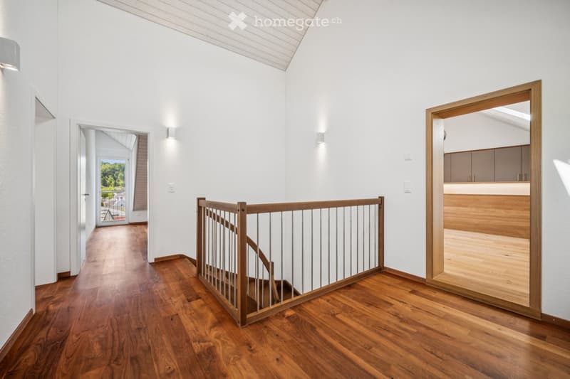 Bright, modern hallway with wooden flooring, white walls, and a wooden staircase railing leading upwards. Natural light streams in from a large window at the end of the corridor.