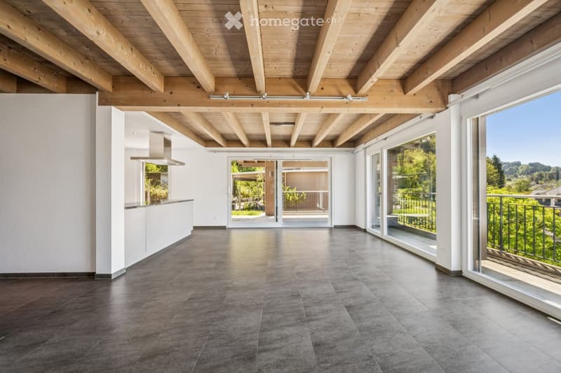 Open-plan space with exposed wooden beams, large windows, and sliding glass doors leading to a balcony. The room features a modern kitchen counter and a sleek range hood.