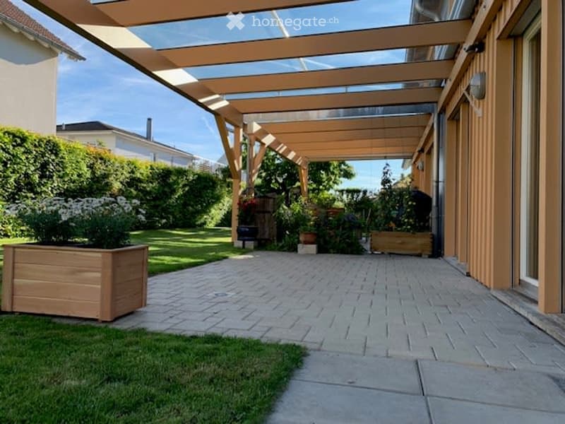 A covered patio area with wooden beams, brick pathway, and surrounding greenery including potted plants and a wooden planter box.