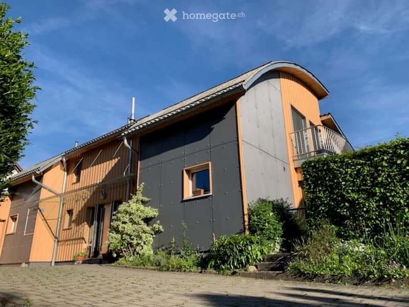 Modern two-story house with a combination of orange and grey exterior walls, large windows, and a small balcony on the upper level.