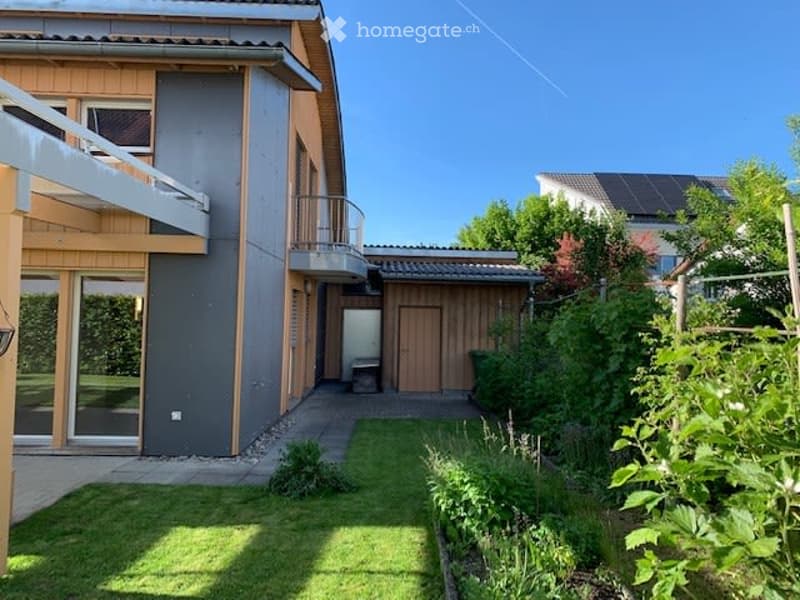 Modern two-story house with a balcony, surrounded by a well-maintained garden with grass and plants.