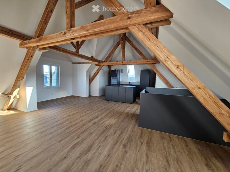 An attic space with exposed wooden beams, wooden flooring, and modern grey cabinets. The room is empty, with no furniture or personal items visible.