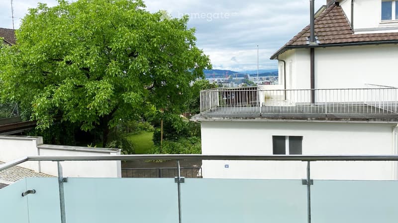 Balcony with metal railing overlooking a tree-lined area and a white building with a rooftop terrace.