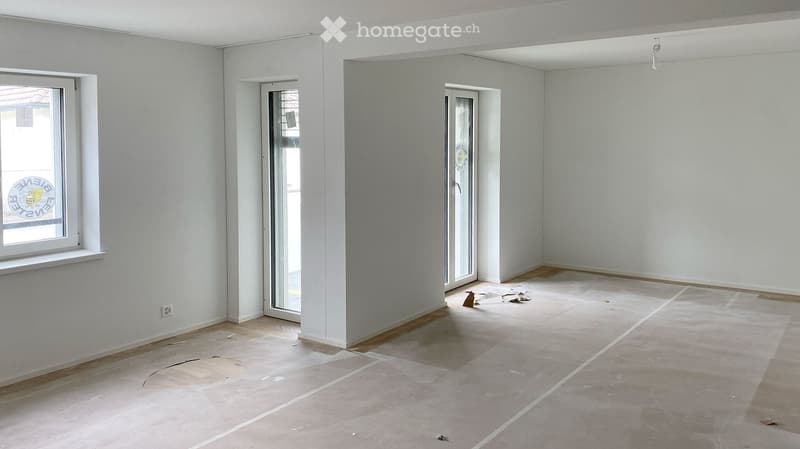 Empty room with light grey walls, wooden baseboards, and unfinished concrete floor. Features include a window with a white frame, a glass door leading outside, and a small electrical outlet on the wall.