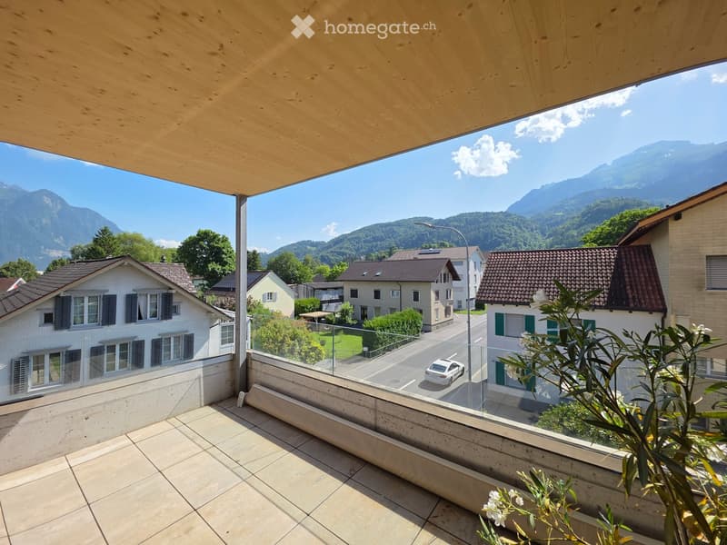 Balcony with wooden ceiling, tiled floor, glass railing, overlooking a street with houses and mountains in the background.