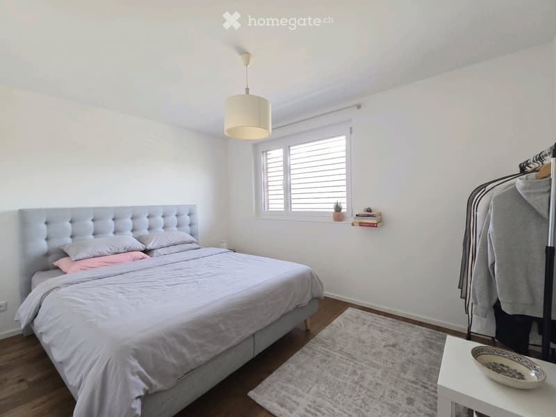 Modern bedroom with a grey upholstered bed, white bedding, wooden floor, and a hanging pendant light. A small nightstand with a lamp sits beside the bed, and a clothing rack with folded clothes is visible.