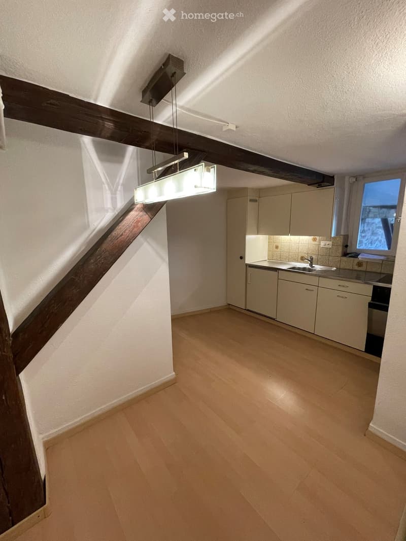 Empty room with wooden flooring, white walls, and a small kitchen area with cabinets and a sink.
