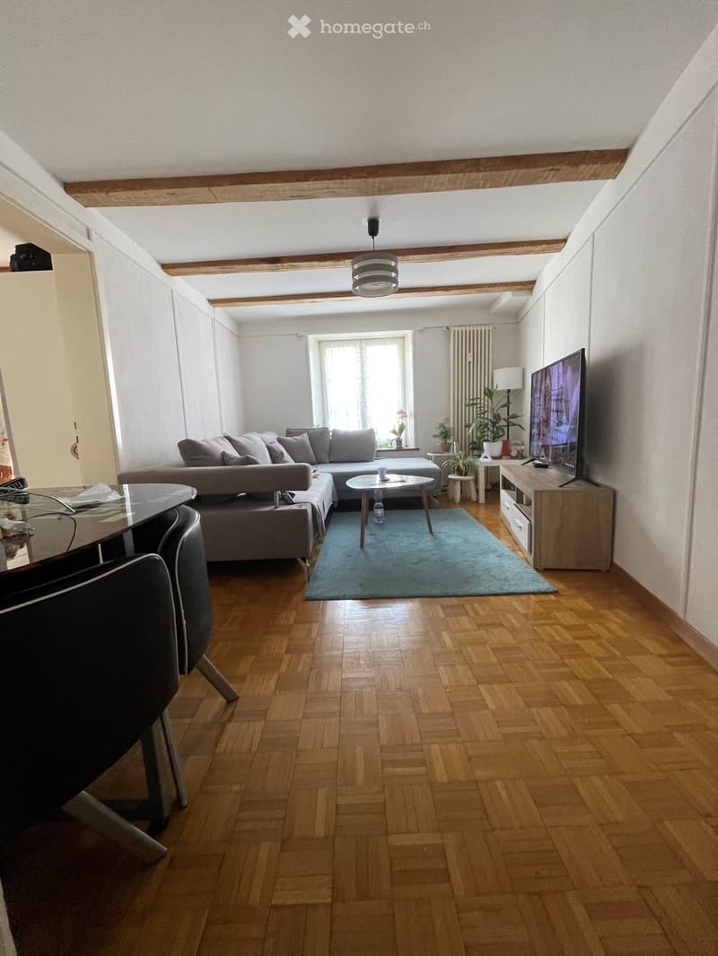 Living room with wooden flooring, exposed wooden beams, a grey sectional sofa, a coffee table, a TV stand, and a dining area with a glass table and chairs.