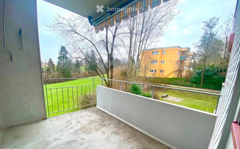 Balcony with metal railing, concrete floor, and striped awning, overlooking a green lawn and residential building.
