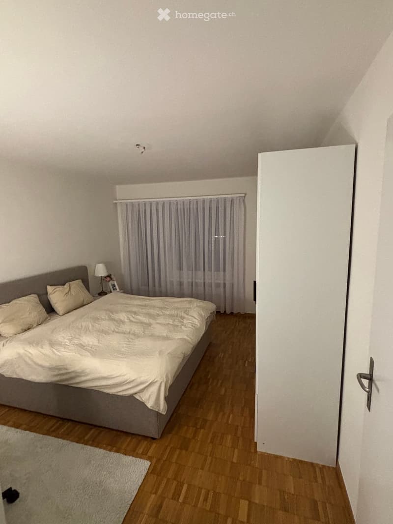 Bedroom with a double bed, nightstand, lamp, and curtains. Wooden flooring and white walls.