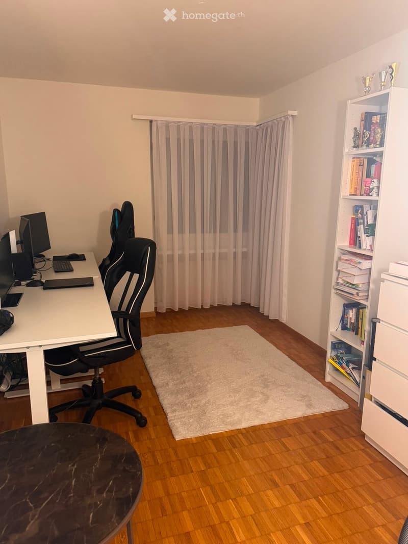 A well-lit room with wooden flooring, a white desk, a black gaming chair, a bookshelf filled with books, and a small white rug.
