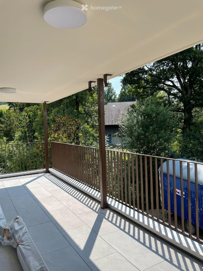 Balcony with a modern design, featuring a tiled floor, wooden railing, and a covered roof supported by metal poles. A portable toilet is visible on the right side.