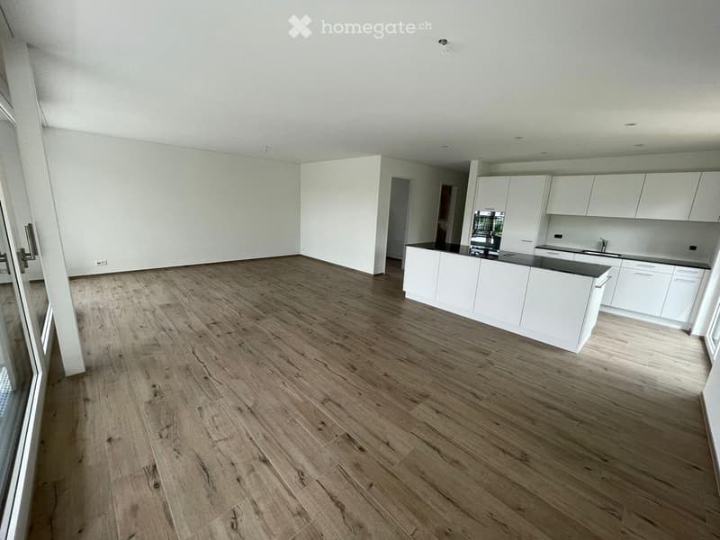 Empty room with light wooden flooring and white walls, featuring a white kitchen area with cabinets and countertops in the background.