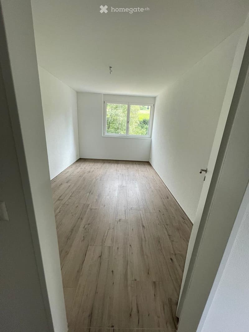 Empty hallway with light wooden flooring and white walls, featuring a window at the far end and a door on the right side.