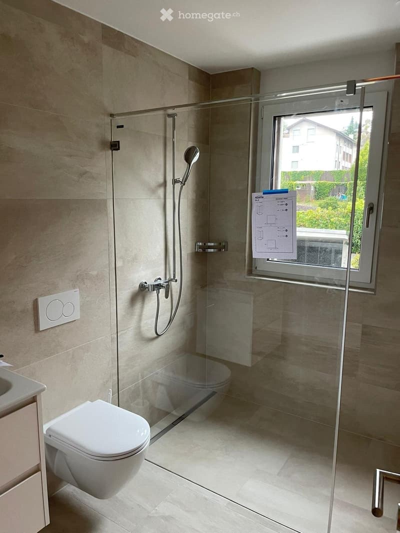 Modern bathroom with a toilet, glass-enclosed shower, and a window offering a view of the outside. The walls and floor are tiled in a neutral color scheme.