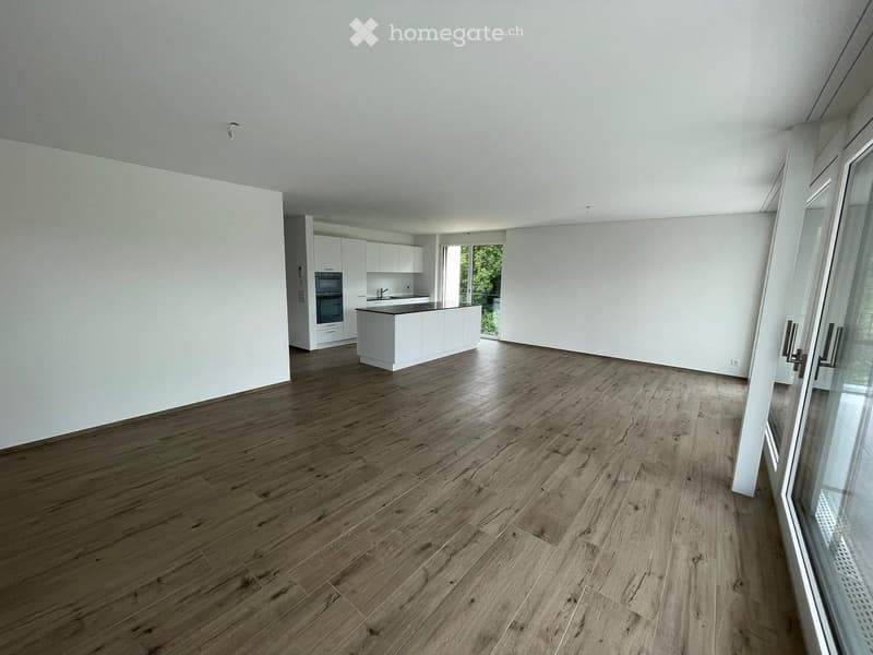 Open-plan space with light wooden flooring, white kitchen cabinets, and a countertop, featuring large windows and a modern design.