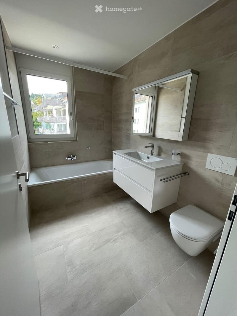 Modern bathroom with a bathtub, sink, toilet, and mirror. The room features light grey tiles and a large window providing natural light.