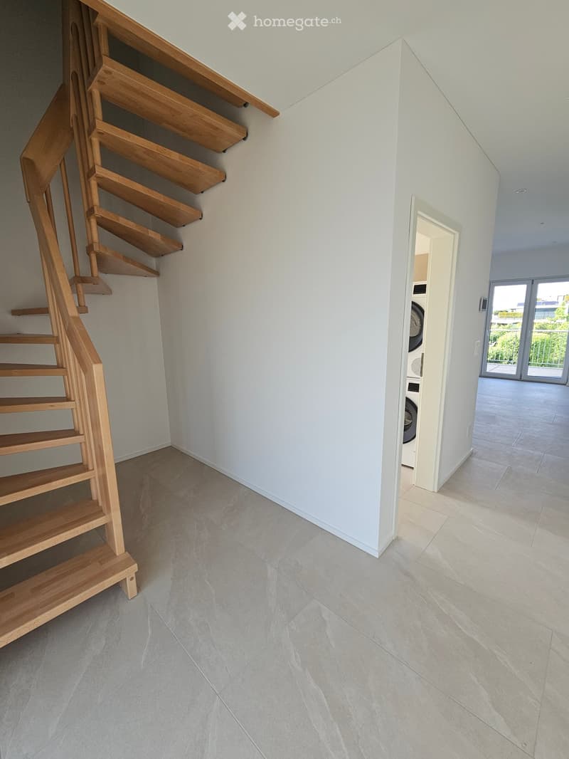 Modern hallway with light-colored tiled flooring, white walls, and a wooden staircase leading upwards. A doorway leads to a room with a washing machine visible.