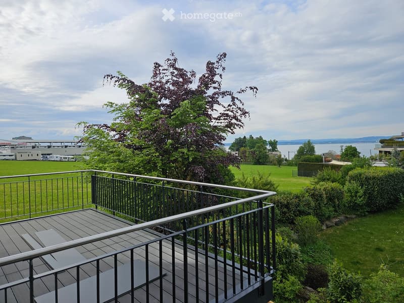 Balcony with wooden decking and metal railing, overlooking a green lawn, trees, and a body of water in the distance.