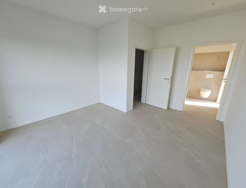 Empty room with light grey tiled flooring and white walls, featuring an open doorway to a bathroom with a toilet visible.
