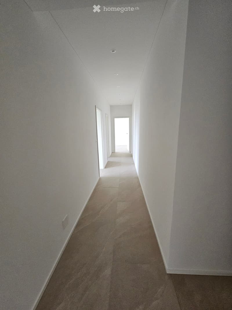 Empty hallway with beige tiled floor and white walls, featuring recessed ceiling lights and a door at the end.