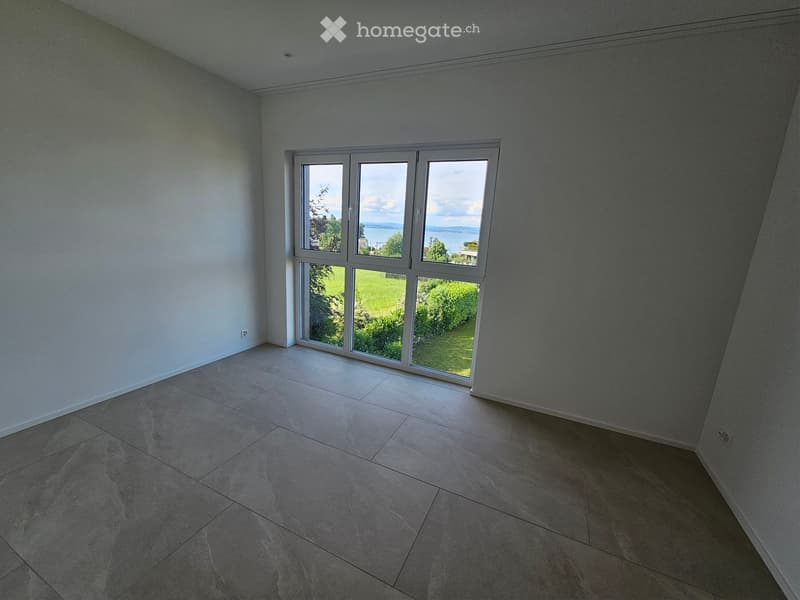 Empty room with tiled floor and large windows offering a view of a green area and water.